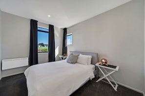 A modern hotel room with a large bed, a bedside table with a lamp, a window with curtains, and a view of the outside. - Modern CBD retreat with parking and courtyard (Christchurch)