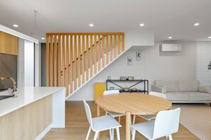Cuina moderna amb una escala de fusta, zona de menjador amb taula rodona i saló amb sofà.
