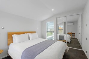 A modern bedroom with a large bed, a wooden headboard, a bedside table, a mirror, and a view of the outdoors. - Modern Town House in the Heart of Merivale (Christchurch)