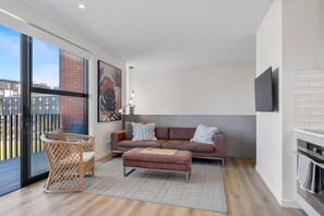 Ein modernes Wohnzimmer mit einer Couch, einem kleinen Tisch und einem Korbstuhl. Es gibt ein großes Fenster und einen Kamin.