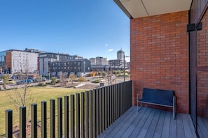 Ein Balkon mit Holzboden und einer Metallbank, mit Blick auf eine Stadtlandschaft mit Gebäuden und einem Parkplatz.