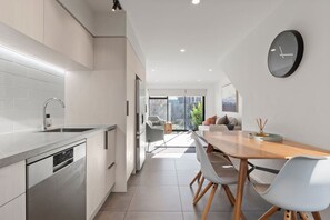 A modern kitchen with a dishwasher, a dining area with a wooden table and chairs, and a large window providing a view of the outdoors. - City Escape | Free Parking + Flexible Beds (Christchurch)