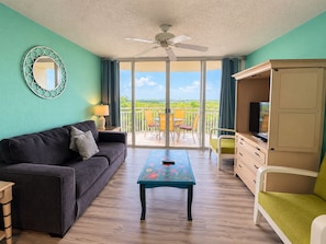 A room with a sofa, a table, a TV stand, and a balcony with chairs. - The Curaçao | Key West Condo with Pool, Balcony, & Sunset Views (Key West)