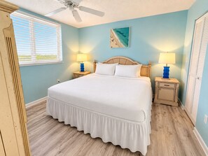 A bedroom with a bed, two bedside tables with lamps, a ceiling fan, a window with blinds, and a wall-mounted picture. - The Curaçao | Key West Condo with Pool, Balcony, & Sunset Views (Key West)
