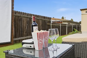 A glass table with a bottle of wine, popcorn containers, and wine glasses. - Outdoor Movie Theater & Fire Pit, Enclosed Yard (La Mesa)