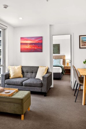 A modern hotel room with a grey sofa, a green ottoman, a bed with a green headboard, a wooden side table, and a painting of a sunset. - 2BR Apartment Near Sky Tower Zodiak Stays (Auckland)