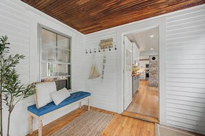 A room with a wooden ceiling, a bench with cushions, a hanging plant, and a view into the kitchen. - Lake Sunapee Swim & Ski Retreat (Newbury)