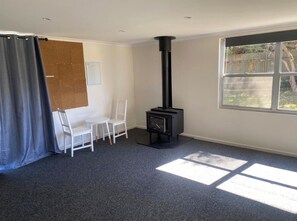 A room with a black wood-burning stove, a white chair, a table, and a large window with a view of greenery. - Wendale Shores on Cave St - minutes to the beach (Goolwa Beach)
