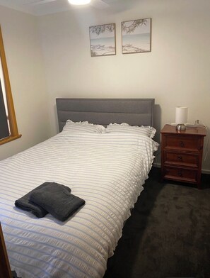 A neatly made bed with a striped comforter and a folded towel, a wooden nightstand with a lamp, and two framed pictures on the wall. - Wendale Shores on Cave St - minutes to the beach (Goolwa Beach)