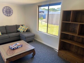 A living room with a sofa, a coffee table with a board game, a clock, and a large window with a view of a backyard. - Wendale Shores on Cave St - minutes to the beach (Goolwa Beach)