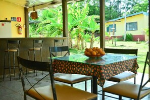An outdoor seating area with a table and chairs, a fire extinguisher on the wall, and a view of a yellow building and greenery. - Suíte Acon. na Natureza - RepousaAquiÓ (Olímpia)