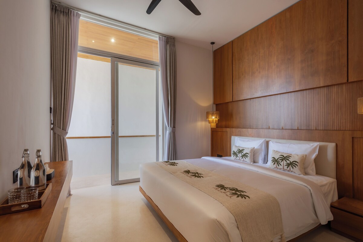 A hotel room with a large bed, a wooden headboard, a nightstand with a lamp, and a sliding glass door leading to a balcony.