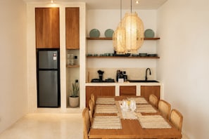A compact kitchen with wooden cabinets, a built-in refrigerator, a hanging light fixture, and a dining area with a wooden table and chairs. - Dimora Bali Resort (Badung)