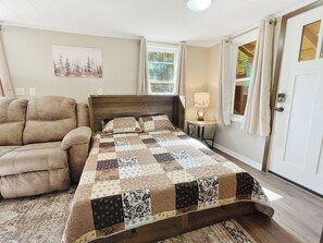 A bedroom with a bed, a chair, a bedside table with a lamp, and a window with curtains. - 2BR Mercer Trailside Retreat near Turtle Flambeau with WiFi, kitchen & deck (Mercer)