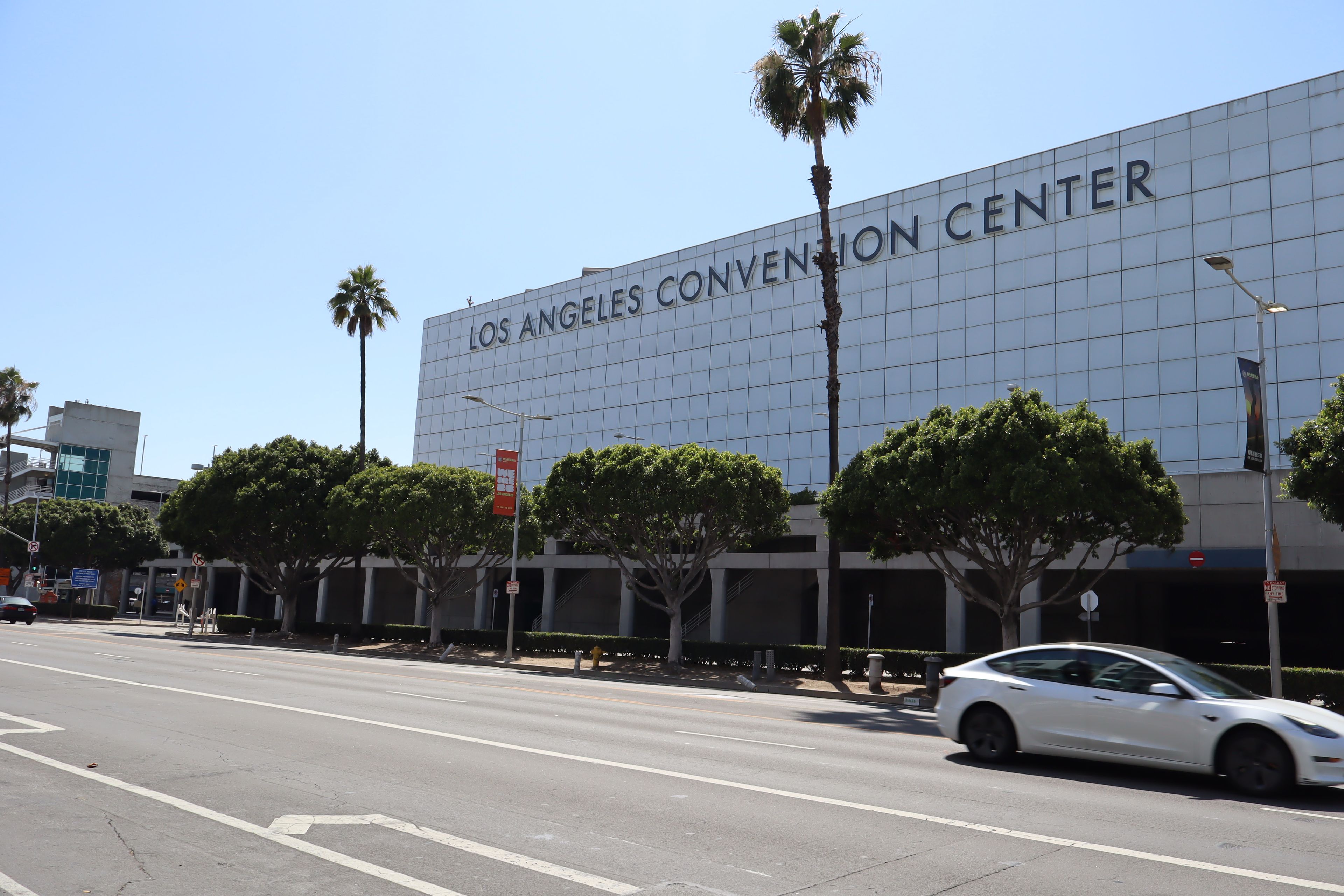 The exterior of the Los Angeles Convention Center with a modern glass facade, palm trees, and a white car parked on the street.