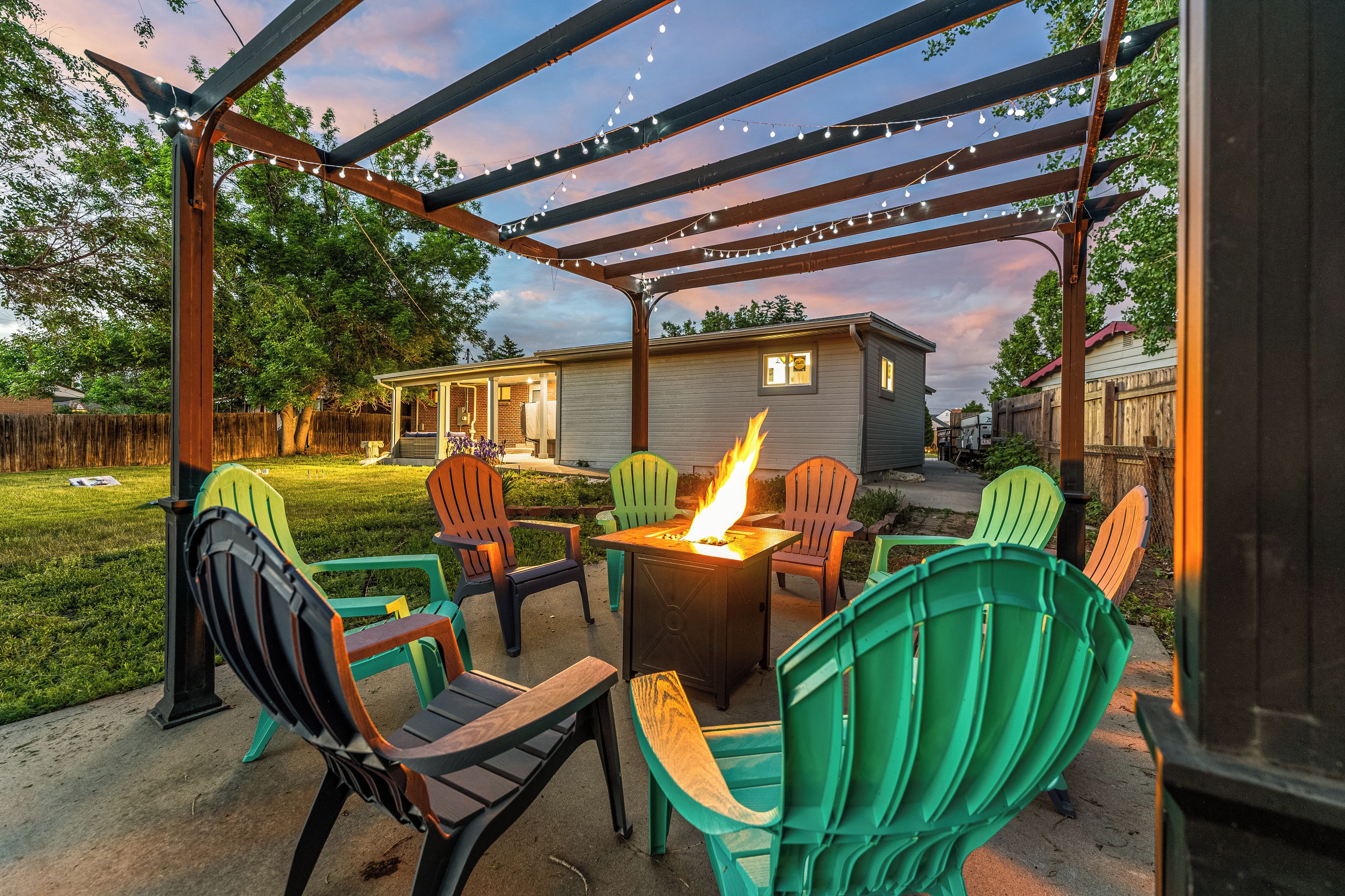 An outdoor patio with a fire feature, green chairs, and a pergola with string lights.