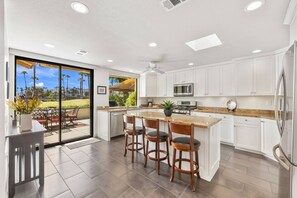 A modern kitchen with white cabinets, a central island with bar stools, and a view of a landscaped area through glass doors. - Gorgeous Monterey Country Club Condo with Fairway Views, Covered Patio & Community Pool (Palm Desert)