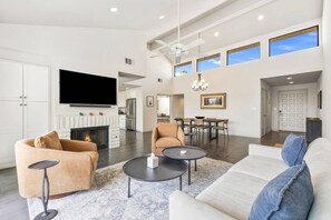 A spacious living room with a fireplace, a television, and a dining area with a chandelier. - Gorgeous Monterey Country Club Condo with Fairway Views, Covered Patio & Community Pool (Palm Desert)
