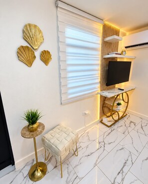 A modern living room with a television, a small round table with a plant, a white cushioned bench, and decorative shells on the wall.