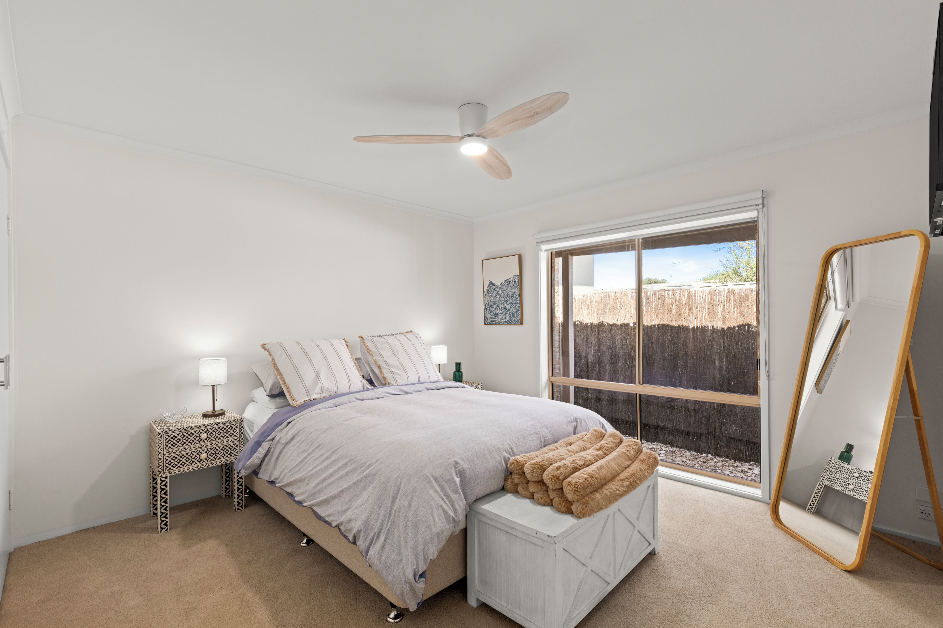 A bedroom with a bed, a bedside table, a lamp, a ceiling fan, a window, and a mirror.