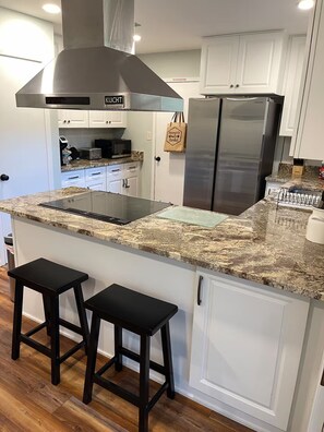 A kitchen with a stainless steel range hood, a double-door refrigerator, granite countertops, and two black bar stools.