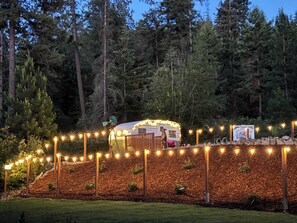 Ein gut beleuchteter Wohnmobilplatz, umgeben von einem Holzzaun und Lichterketten, mit einem Wald im Hintergrund.