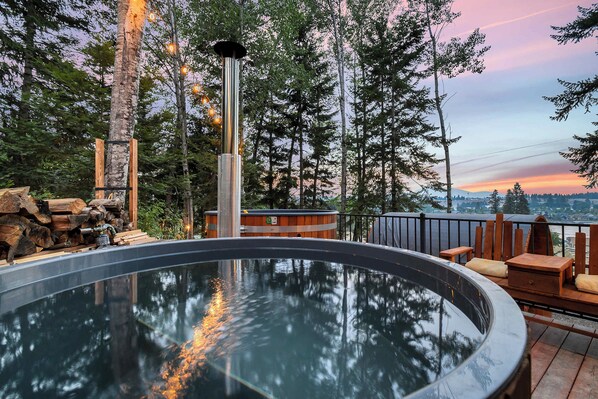 A hot tub surrounded by wooden logs, a metal structure, and a view of a city skyline at sunset.