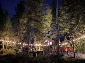 A cabin surrounded by tall trees at night with string lights.