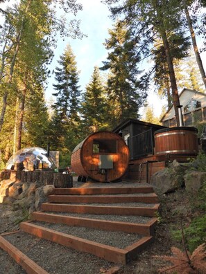 A wooden cabin with a large barrel-like structure, surrounded by trees and a house in the background.