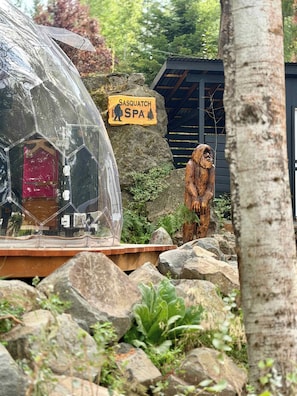 A transparent geodesic dome structure with a red door, a sign for Sasquatch Spa, and a wooden sculpture of a sasquatch. - Sasquatch Lodge: Private Pool, Spa & Stargazing Dome (Coeur d'Alene)