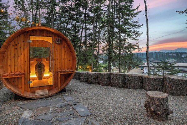 A wooden sauna with a glass door, surrounded by a gravel path and trees, with a view of a cityscape in the background.