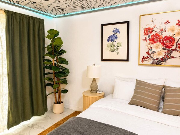 A hotel room with a bed, a bedside table with a lamp, a large potted plant, and two framed floral artworks on the wall. - The Cozy Koi Walk to the CDA Resort With Private Porch (Coeur d'Alene)