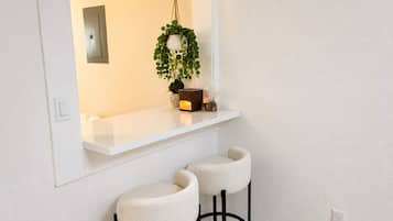 A clean, modern interior with a white wall, a white shelf, two white barstools, a hanging plant, and a small decorative item.