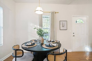 A dining area with a round table, four chairs, a hanging light fixture, a window with curtains, and a door with a picture on the wall. - Heart ATL– 2BR/ Monthly Stays /Pet-Friendly (Atlanta)