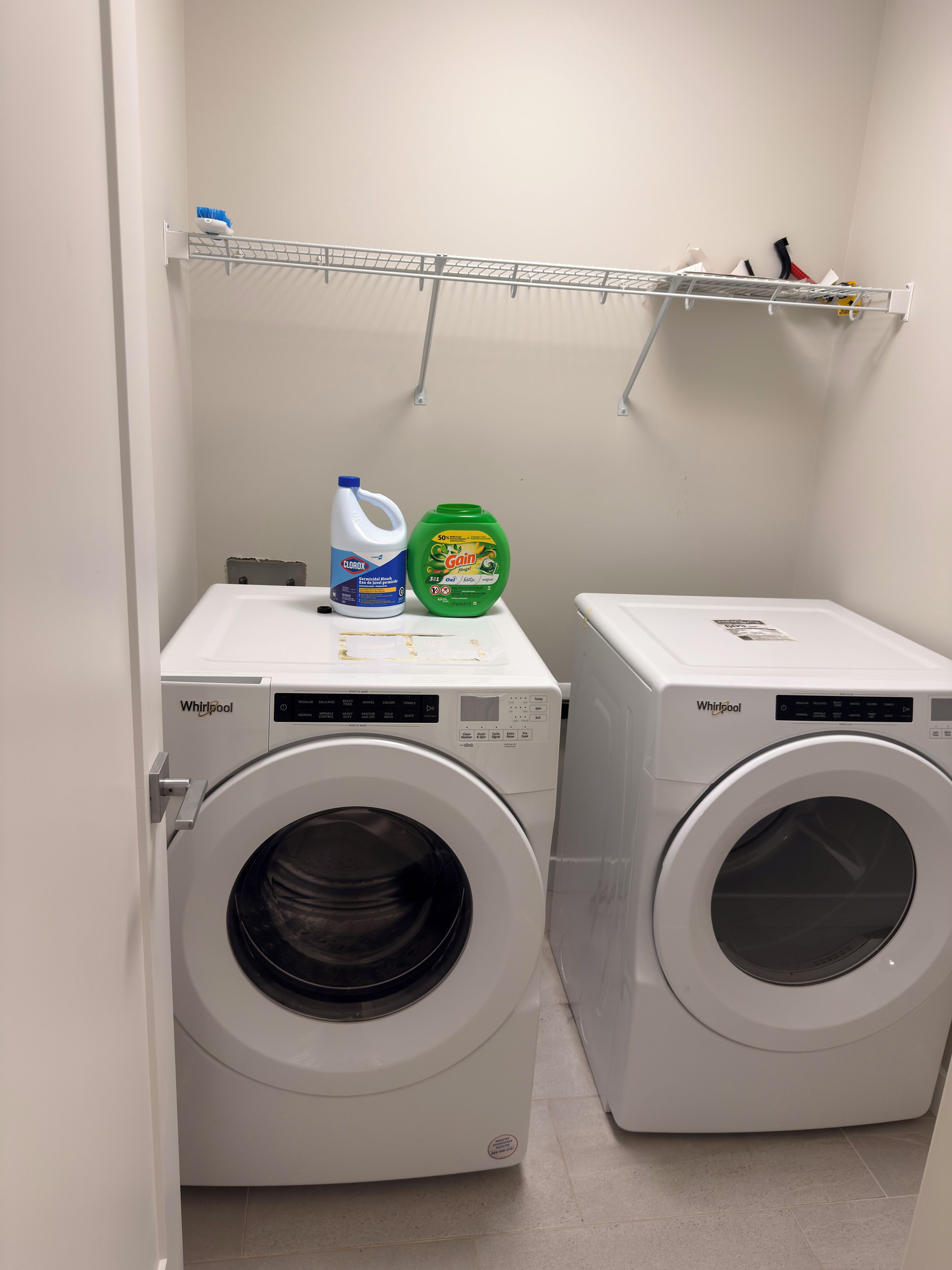 Two Whirlpool washing machines in a laundry room with a shelf above holding cleaning supplies.
