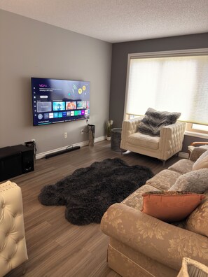 Ein modernes Wohnzimmer mit einem an der Wand montierten Flachbildfernseher, einem Sofa, einem flauschigen Teppich und einem Fenster mit Jalousien.