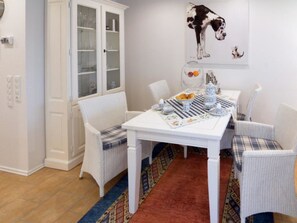 A dining area with a white table set for four, surrounded by wicker chairs, a patterned rug, and a wall-mounted painting of a dog. - Holiday Apartment With 1 Bedroom (Norden)
