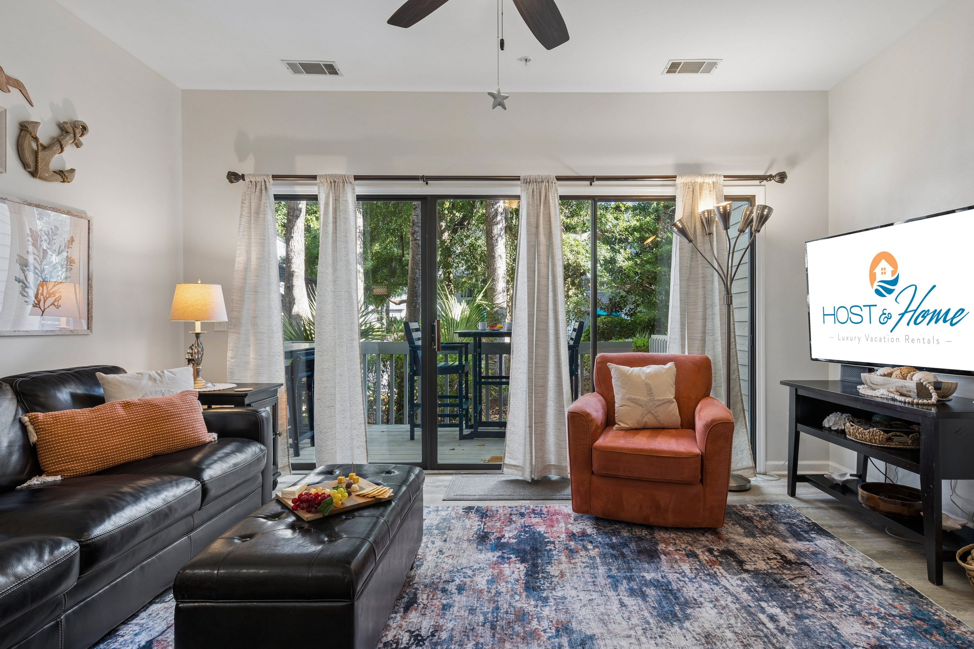 A modern living room with a leather sofa, a brown armchair, a coffee table with fruit, and a large TV screen displaying 'HOST & Home - Luxury Vacation Rentals'.