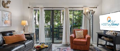 A modern living room with a leather sofa, a brown armchair, a coffee table with fruit, and a large TV screen displaying 'HOST & Home - Luxury Vacation Rentals'.
