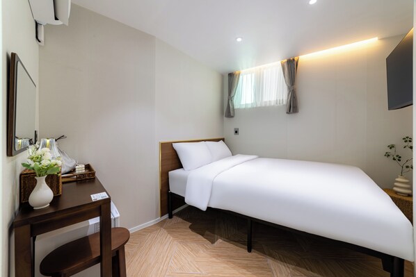 A single bed with a wooden headboard, a bedside table with a vase of flowers, a mirror on the wall, and a window with curtains.