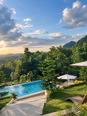 A pool area with a view of a forested hillside, surrounded by greenery and a clear sky.