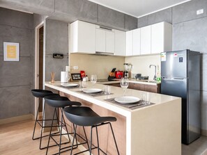 A modern kitchen with a central island, bar stools, a refrigerator, and a dining area. - Stylish Ocean View 2BR • King Bed• Pool & Gym (Barranco)