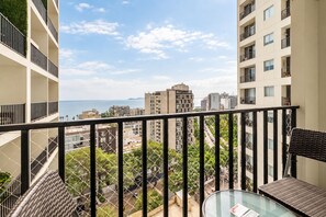 Ein Balkon mit Glastisch, Korbstuhl und Blick auf das Meer und die Stadt.