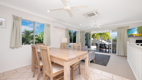 A modern hotel room with a dining area, a sofa, a TV, and a balcony with a view of palm trees.