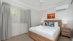 A hotel room with a bed, two bedside tables, a ceiling fan, an air conditioning unit, and a painting of a sunset over the ocean.