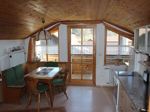 A dining area with a wooden table and chairs, a kitchen with a stove and cabinets, and large windows with curtains. - Holiday Apartment With 2 Bedrooms (Rosshaupten)