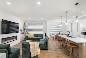 Ein modernes Wohnzimmer mit Kamin, grünen Sofas, einer Essecke mit Barhocker und einer Küche mit Pendelleuchten.