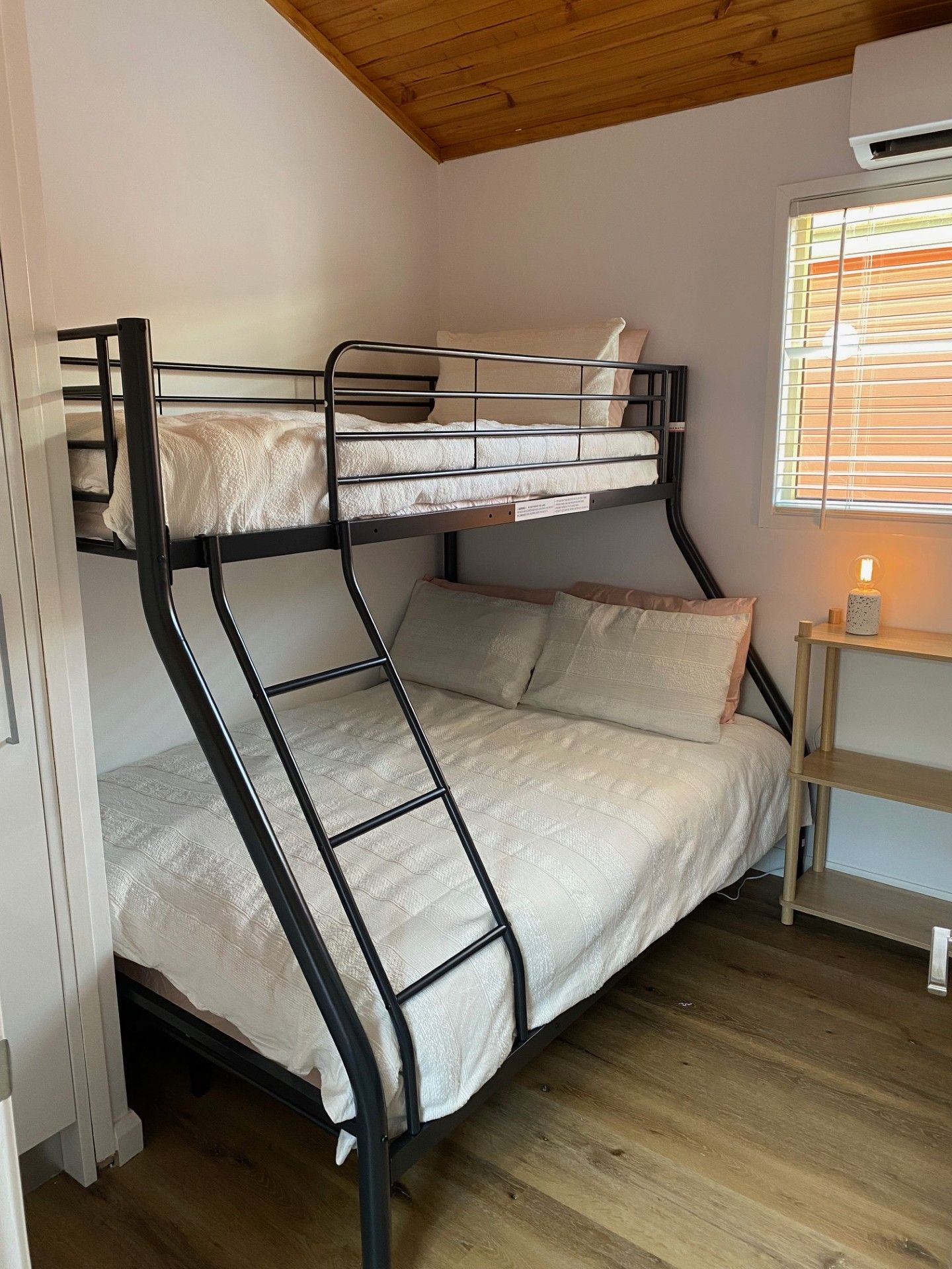 Un lit superposé avec une échelle, une table de chevet avec une lampe et une fenêtre avec des stores.