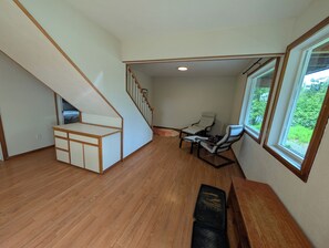 Une pièce spacieuse avec un plancher en bois, un escalier, un coin salon avec des fauteuils et une fenêtre donnant sur un paysage verdoyant.