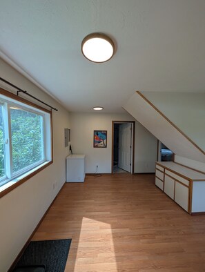 A spacious room with a large window, a ceiling light, a small white cabinet, and a door leading to another room. - Cozy Chalet in the Heart of Girdwood (Anchorage)